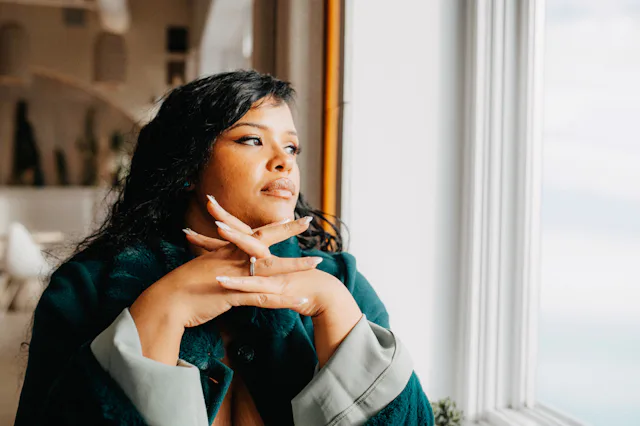 Woman with dark hair and coat resting chin on folded hands, gazing out a window in a softly lit inte...