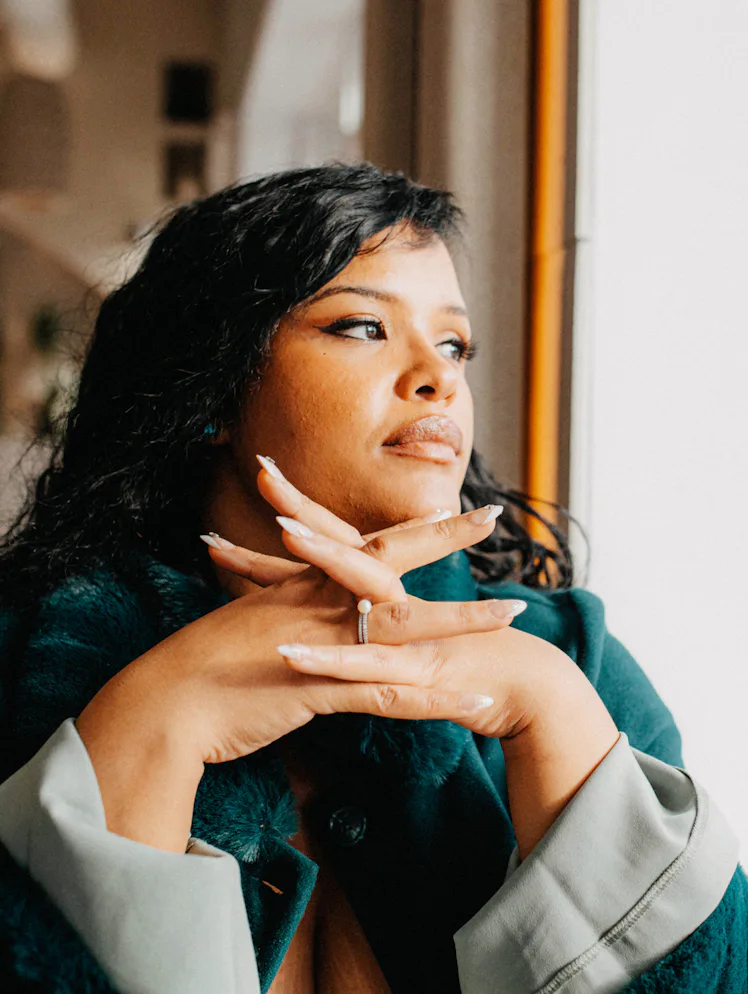 Woman with dark hair and coat resting chin on folded hands, gazing out a window in a softly lit inte...