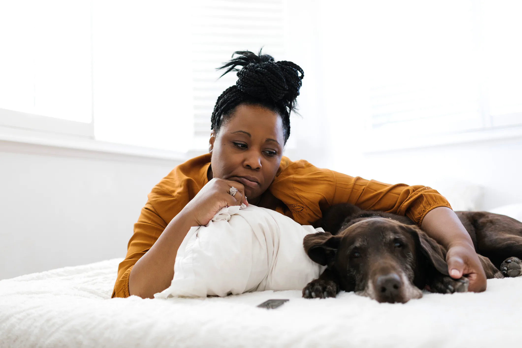 Middle-aged Black woman in a mustard shirt lies on a white bed, resting her head on a pillow and her...
