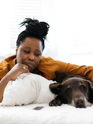 Middle-aged Black woman in a mustard shirt lies on a white bed, resting her head on a pillow and her...