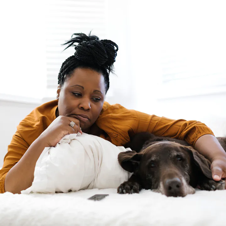 Middle-aged Black woman in a mustard shirt lies on a white bed, resting her head on a pillow and her...
