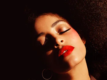 a person with red lipstick and curly hair and prominent pink blush takes in the sun while shadows pl...