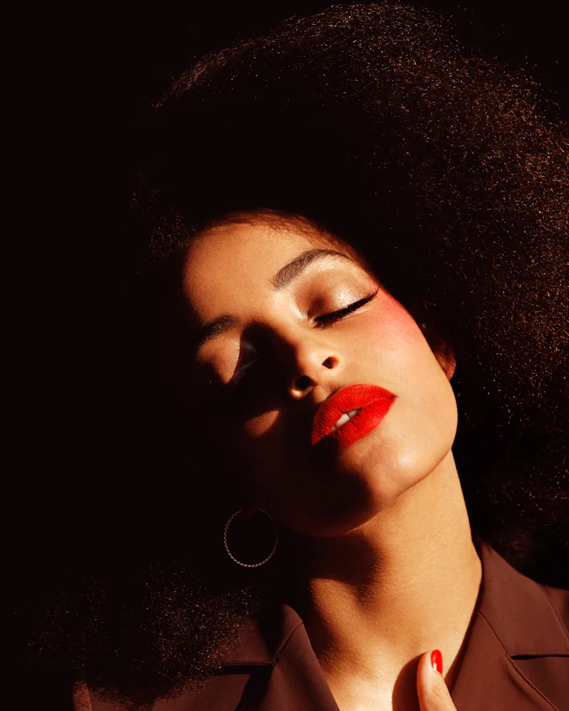 a person with red lipstick and curly hair and prominent pink blush takes in the sun while shadows pl...
