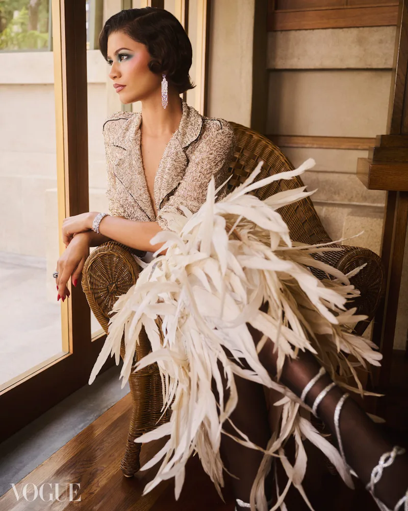 Zendaya sitting in a chair, wearing feathers, with '80s makeup and '20s hair