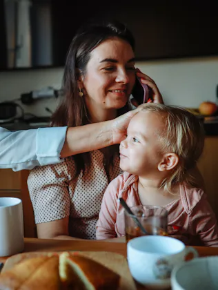 Three generations sit at a kitchen table: an older woman smiling and reaching to touch a toddler’s h...
