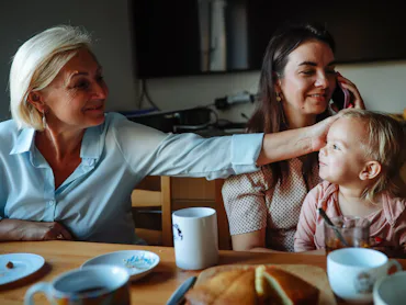 Three generations sit at a kitchen table: an older woman smiling and reaching to touch a toddler’s h...