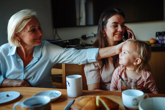 Three generations sit at a kitchen table: an older woman smiling and reaching to touch a toddler’s h...