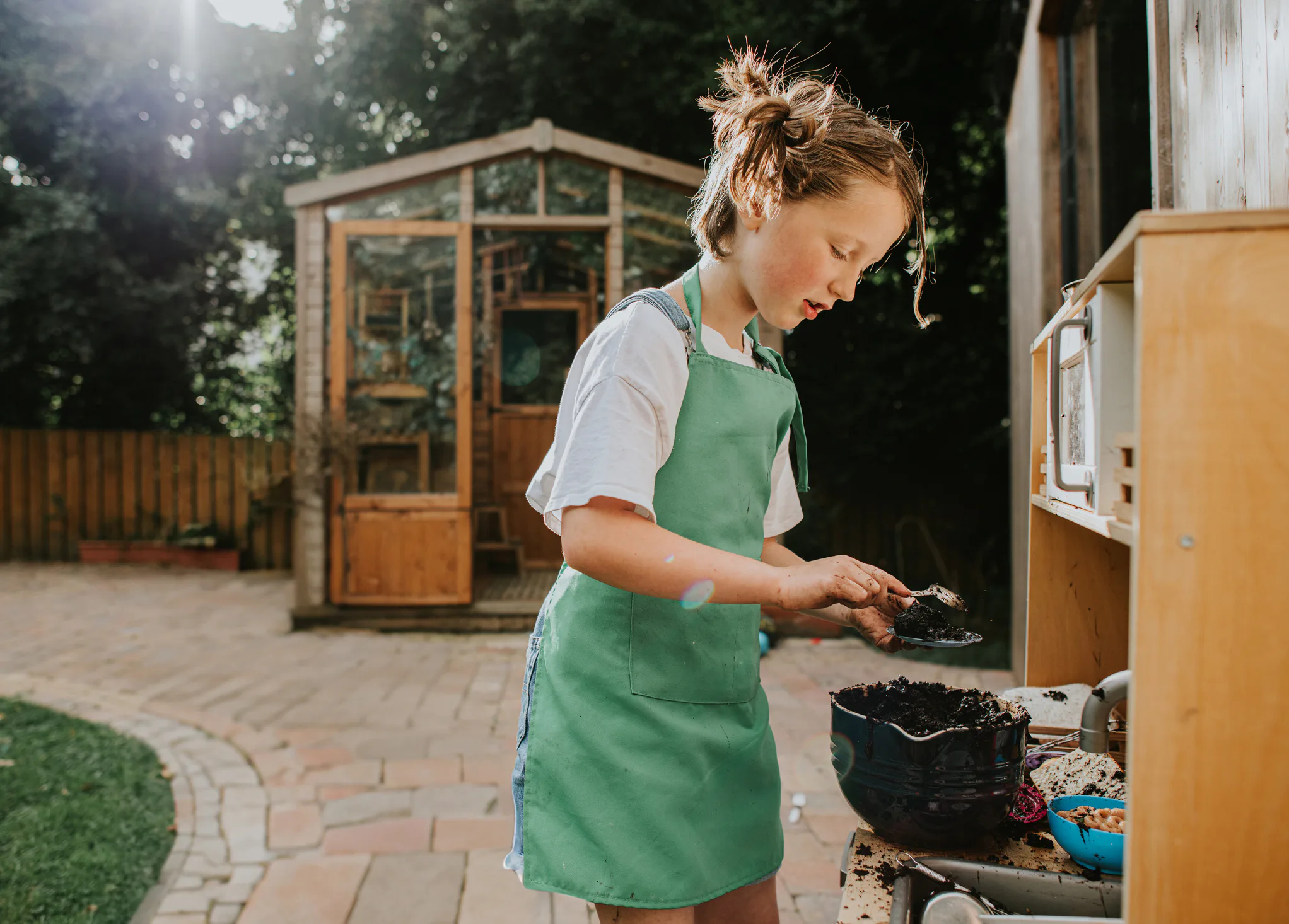 Child in a green apron scooping soil into a small pot at an outdoor play kitchen, with a wooden shed...