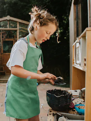 Child in a green apron scooping soil into a small pot at an outdoor play kitchen, with a wooden shed...