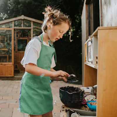 Child in a green apron scooping soil into a small pot at an outdoor play kitchen, with a wooden shed and garden path in the background.