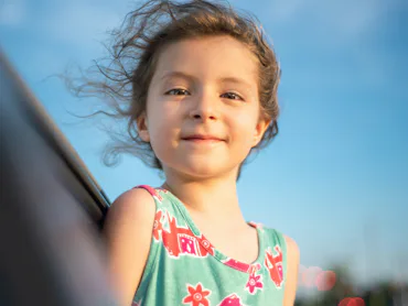 Young girl with windblown hair, wearing a colorful sleeveless dress, smiling slightly against a clea...