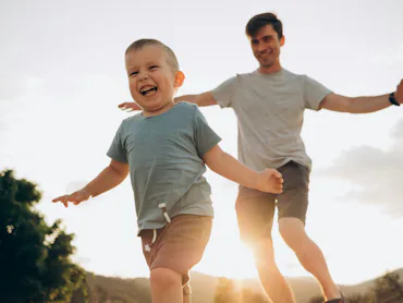 Young boy laughing and running toward camera with outstretched arms as an adult man, likely his fath...