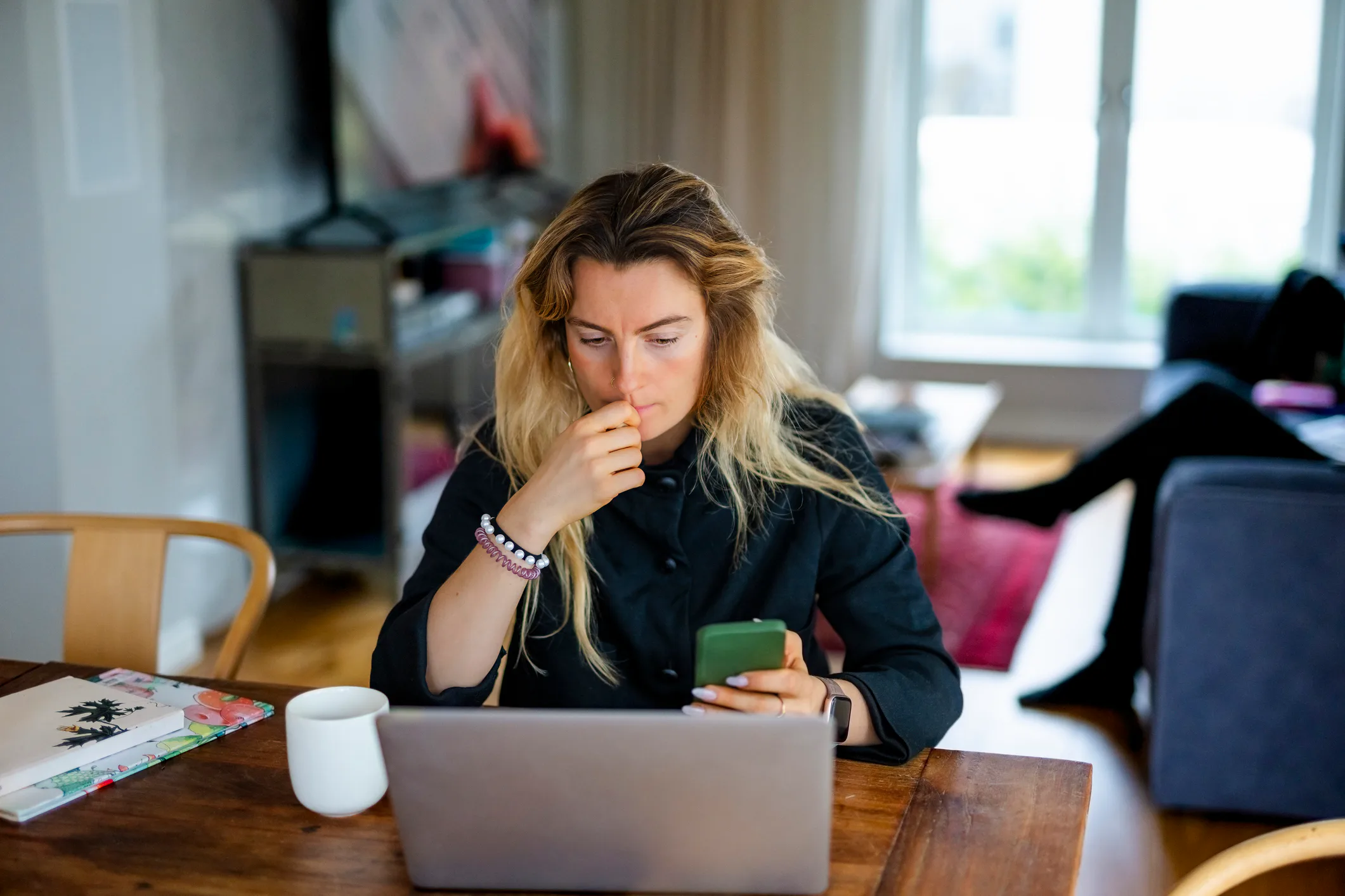 Woman with long hair sits at a wooden table in a living room, looking thoughtful while holding a sma...