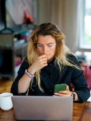 Woman with long hair sits at a wooden table in a living room, looking thoughtful while holding a sma...