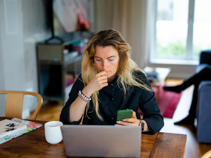 Woman with long hair sits at a wooden table in a living room, looking thoughtful while holding a sma...