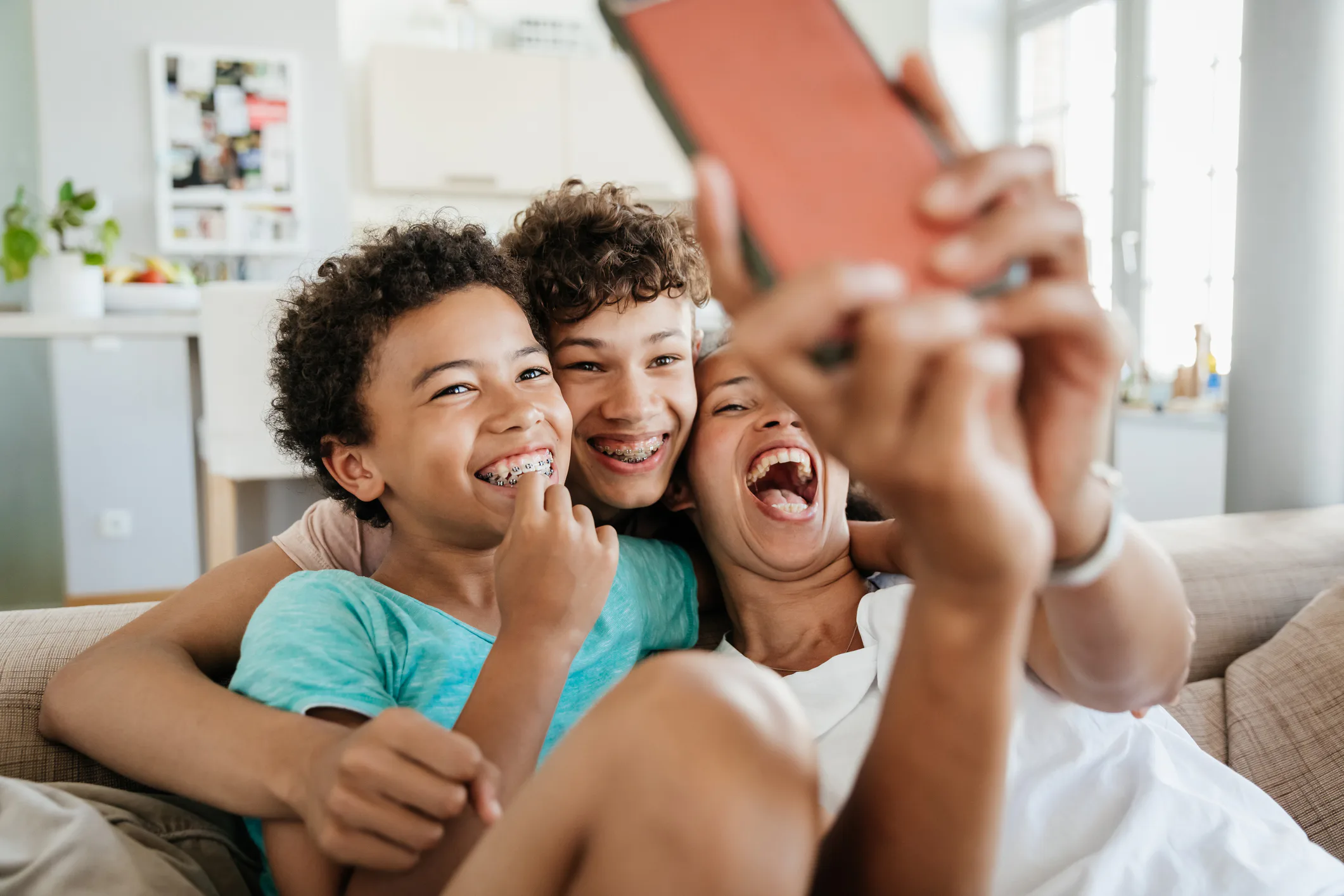 Three smiling siblings with braces sit close on a couch in a bright living room, one holding a phone...