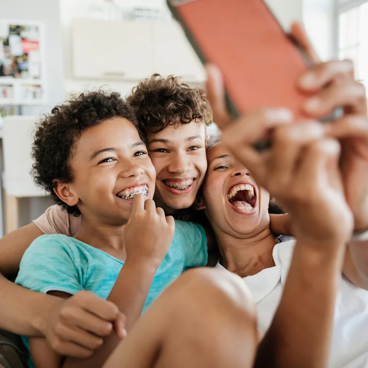 Three smiling siblings with braces sit close on a couch in a bright living room, one holding a phone...