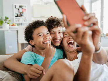 Three smiling children with braces sit closely on a couch, laughing and hugging as one holds a smart...