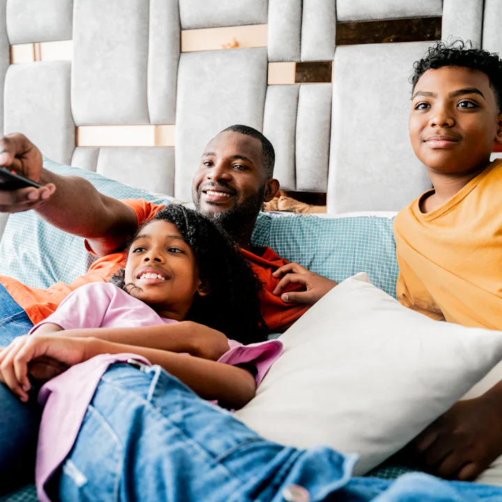 Father and two children reclining on a bed, father pointing a TV remote while kids relax against pil...