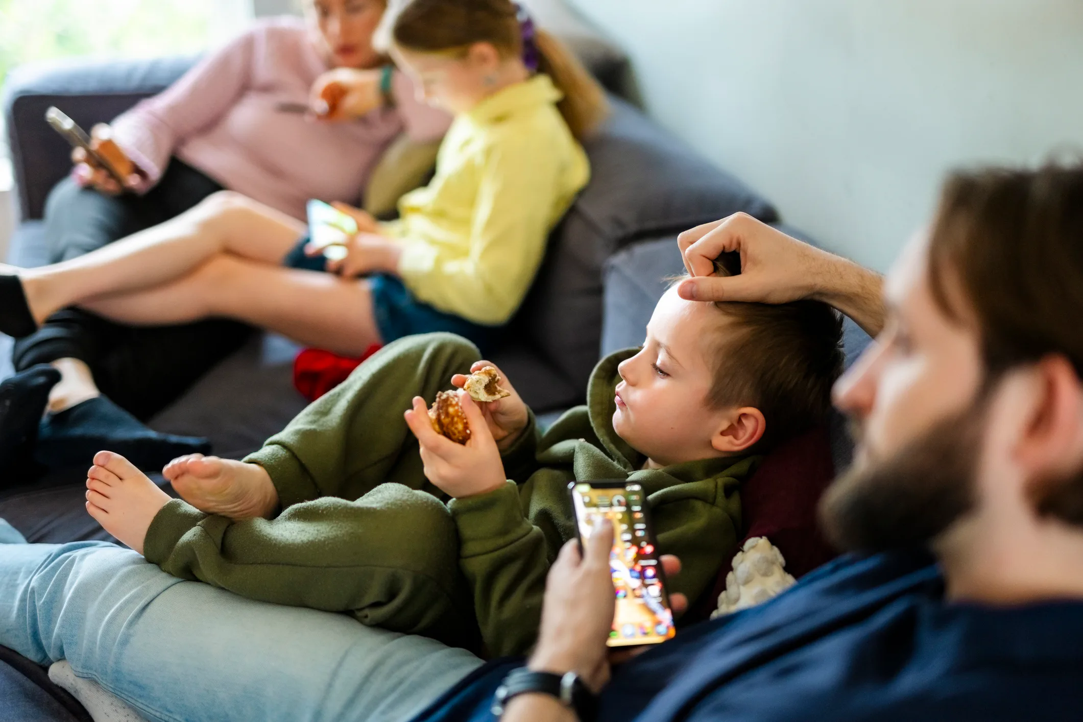 Family members lounging on a couch: two children eating snacks and using tablets while adults relax ...