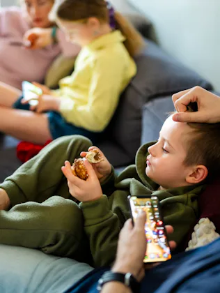 Family members lounging on a couch: two children eating snacks and using tablets while adults relax ...