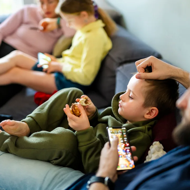 Family members lounging on a couch: two children eating snacks and using tablets while adults relax ...