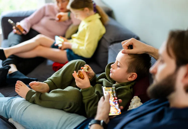 Family members lounging on a couch: two children eating snacks and using tablets while adults relax ...