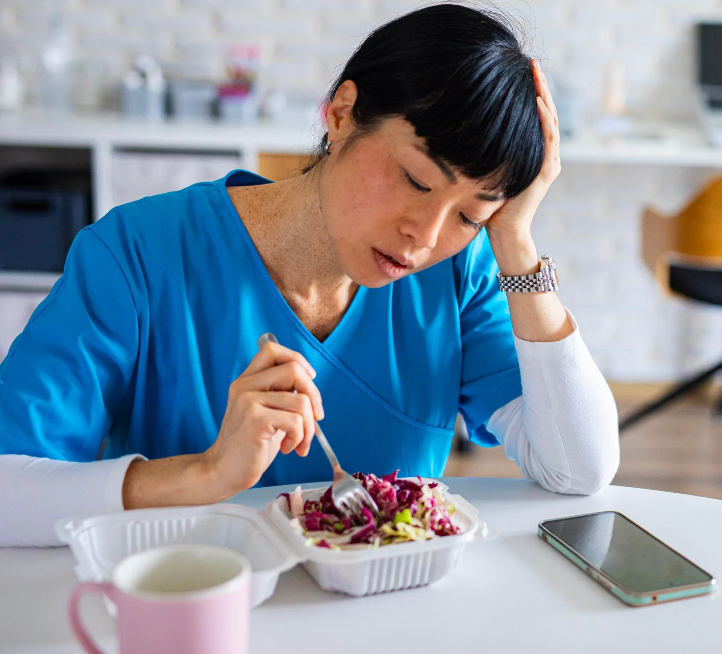 Tired-looking healthcare worker in blue scrubs sits at a table, resting her head while poking a take...