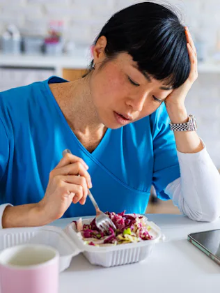 Tired-looking healthcare worker in blue scrubs sits at a table, resting her head while poking a take...