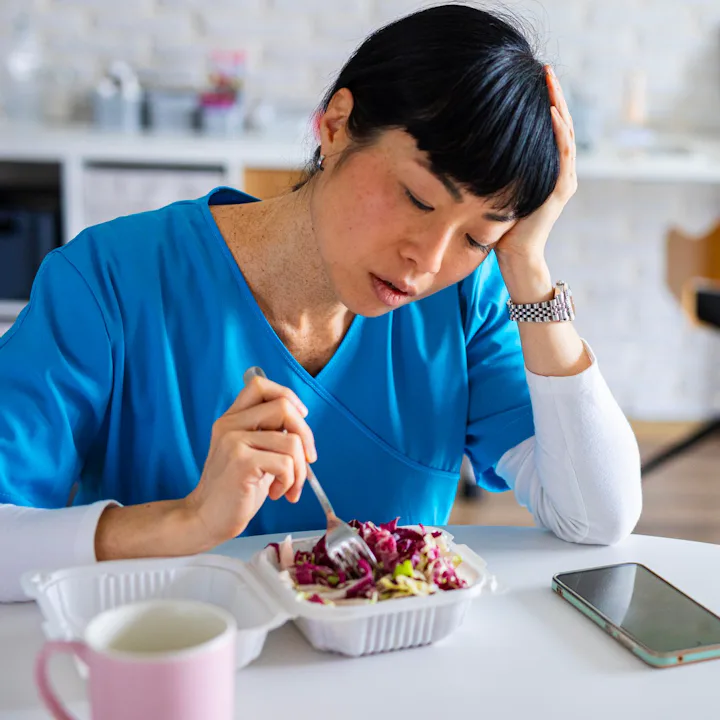 Tired-looking healthcare worker in blue scrubs sits at a table, resting her head while poking a take...