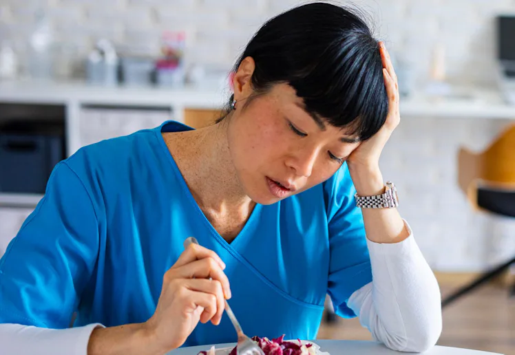 Tired-looking healthcare worker in blue scrubs sits at a table, resting her head while poking a take...