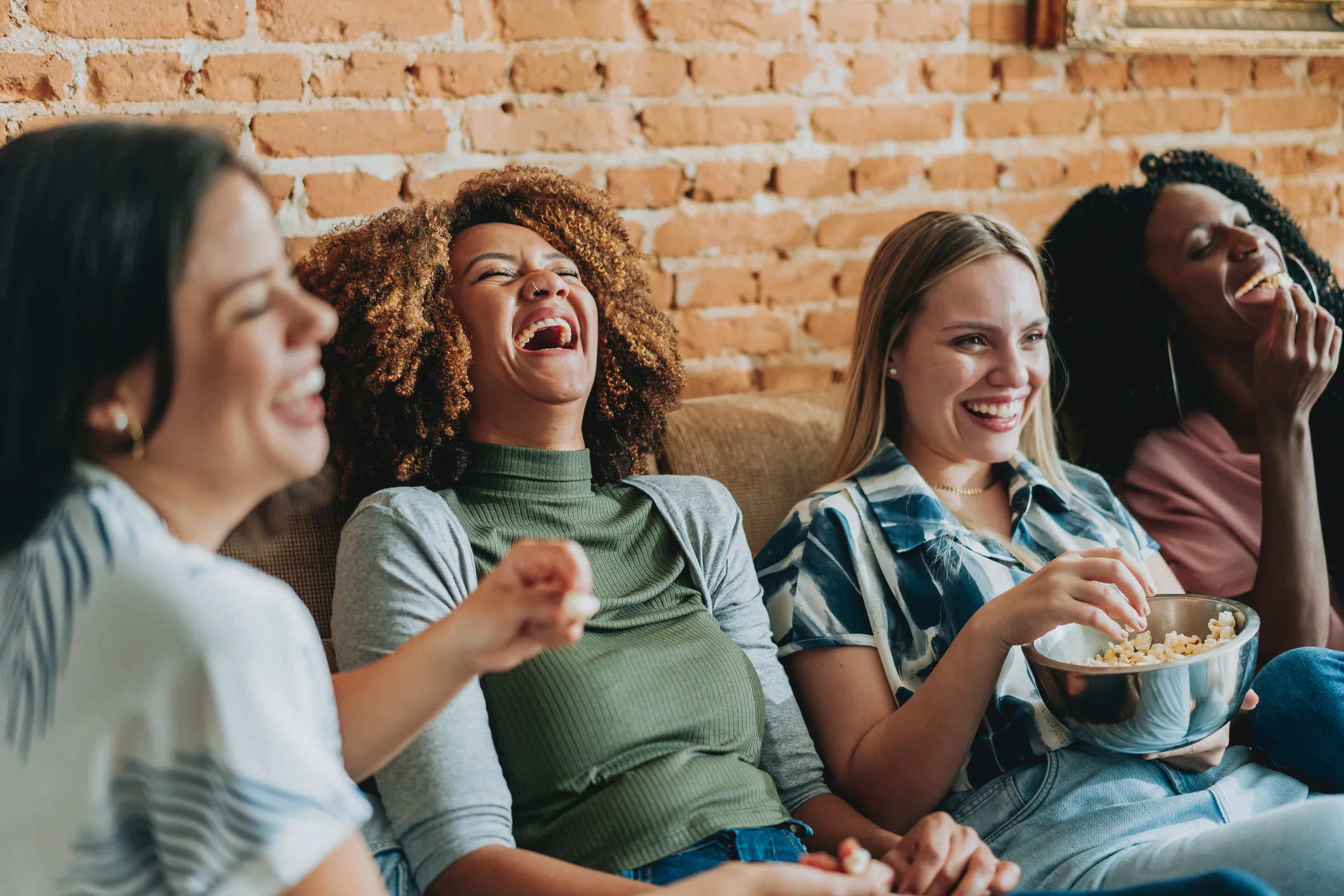 Four women sitting on a couch against a brick wall, laughing together; one holds a bowl of popcorn w...