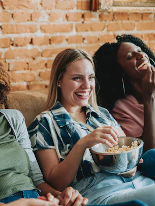 Four women sitting on a couch against a brick wall, laughing together; one holds a bowl of popcorn w...