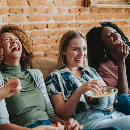 Four women sitting on a couch against a brick wall, laughing together; one holds a bowl of popcorn w...