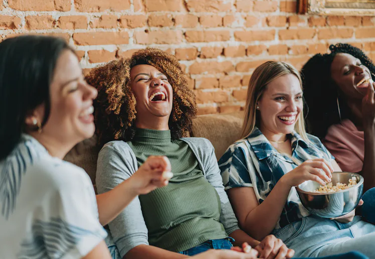 Four women sitting on a couch against a brick wall, laughing together; one holds a bowl of popcorn w...