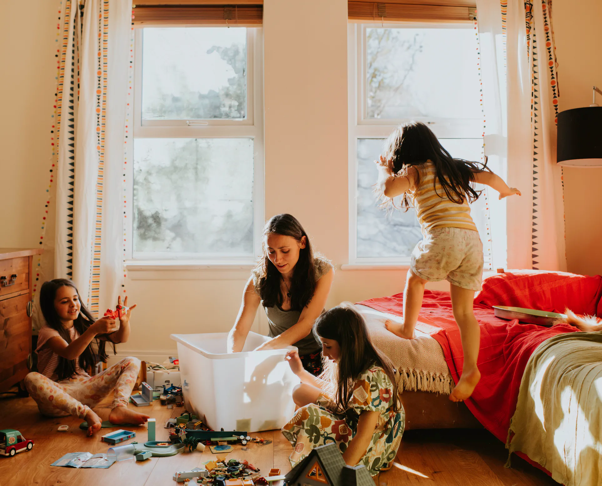 Woman and three girls in a sunlit bedroom: two girls sit on floor playing with toys and a storage bi...