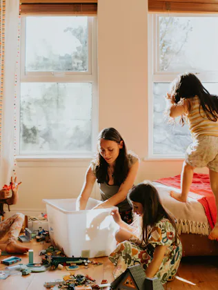 Woman and three girls in a sunlit bedroom: two girls sit on floor playing with toys and a storage bi...