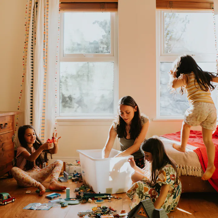 Woman and three girls in a sunlit bedroom: two girls sit on floor playing with toys and a storage bi...
