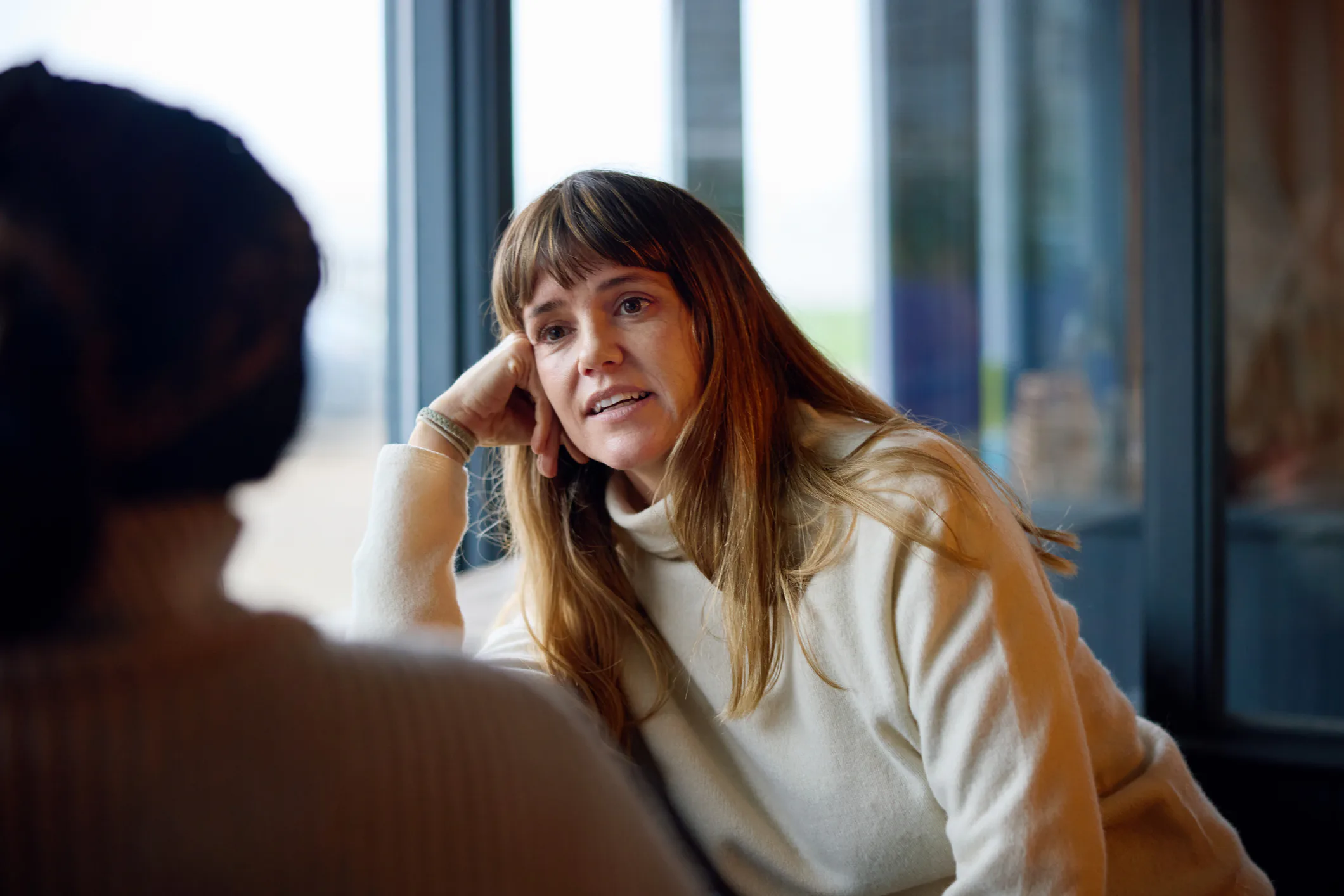 Woman with long brown hair and bangs wearing a cream sweater, leaning on her hand and engaged in con...
