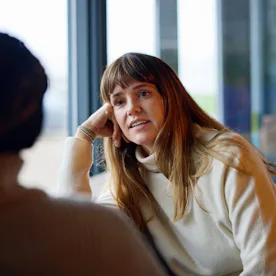 Woman with long brown hair and bangs wearing a cream sweater, leaning on her hand and engaged in con...