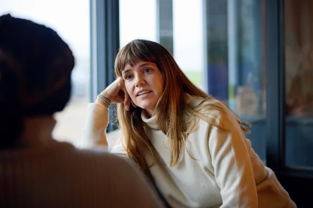 Woman with long brown hair and bangs wearing a cream sweater, leaning on her hand and engaged in con...