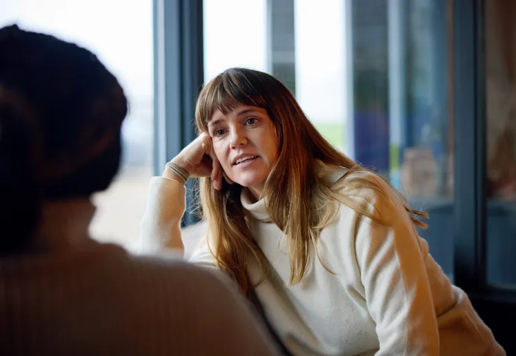 Woman with long brown hair and bangs wearing a cream sweater, leaning on her hand and engaged in con...