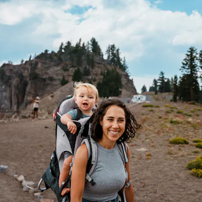 Smiling woman hiking on a rocky trail carries a laughing toddler in a backpack carrier, with pine trees, rugged cliffs and cloudy sky behind them.
