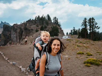 Smiling woman hiking on a rocky trail carries a laughing toddler in a backpack carrier, with pine tr...