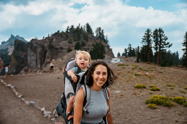 Smiling woman hiking on a rocky trail carries a laughing toddler in a backpack carrier, with pine tr...