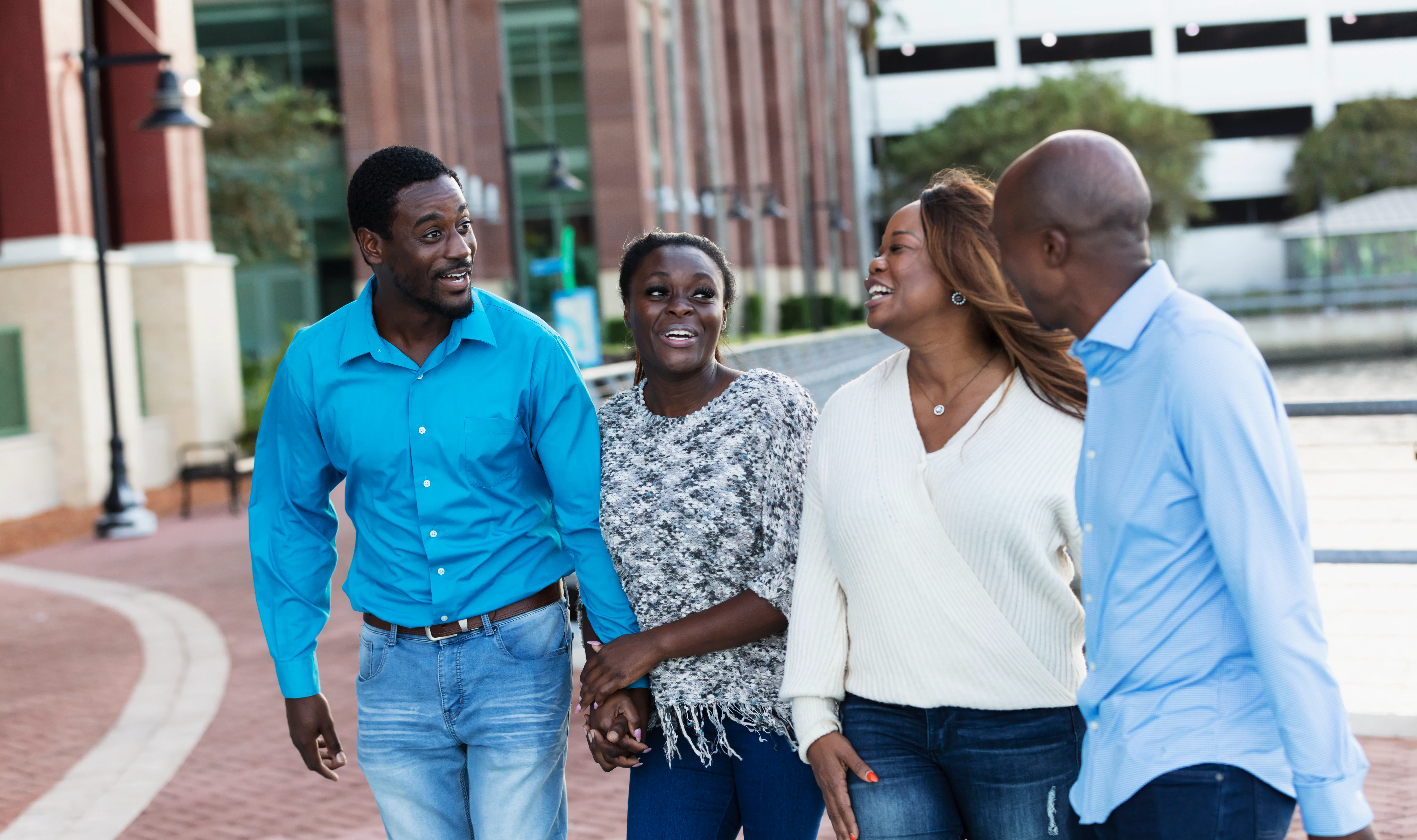 Four adults walk outdoors along a waterfront plaza, smiling and talking; two women and two men hold ...