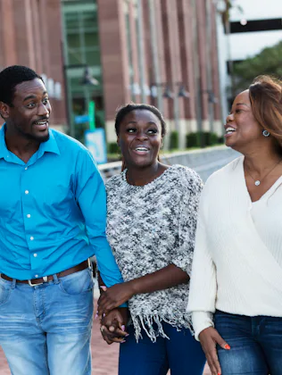 Four adults walk outdoors along a waterfront plaza, smiling and talking; two women and two men hold ...