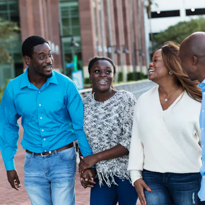 Four adults walk outdoors along a waterfront plaza, smiling and talking; two women and two men hold hands and gesture as they stroll past buildings.