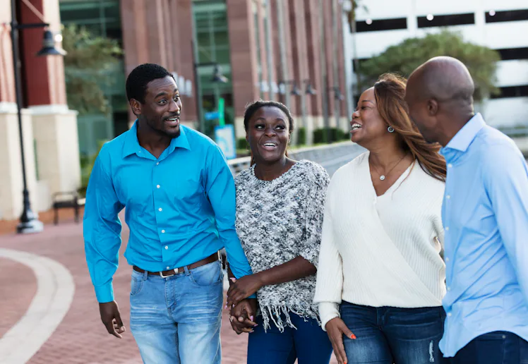 Four adults walk outdoors along a waterfront plaza, smiling and talking; two women and two men hold ...