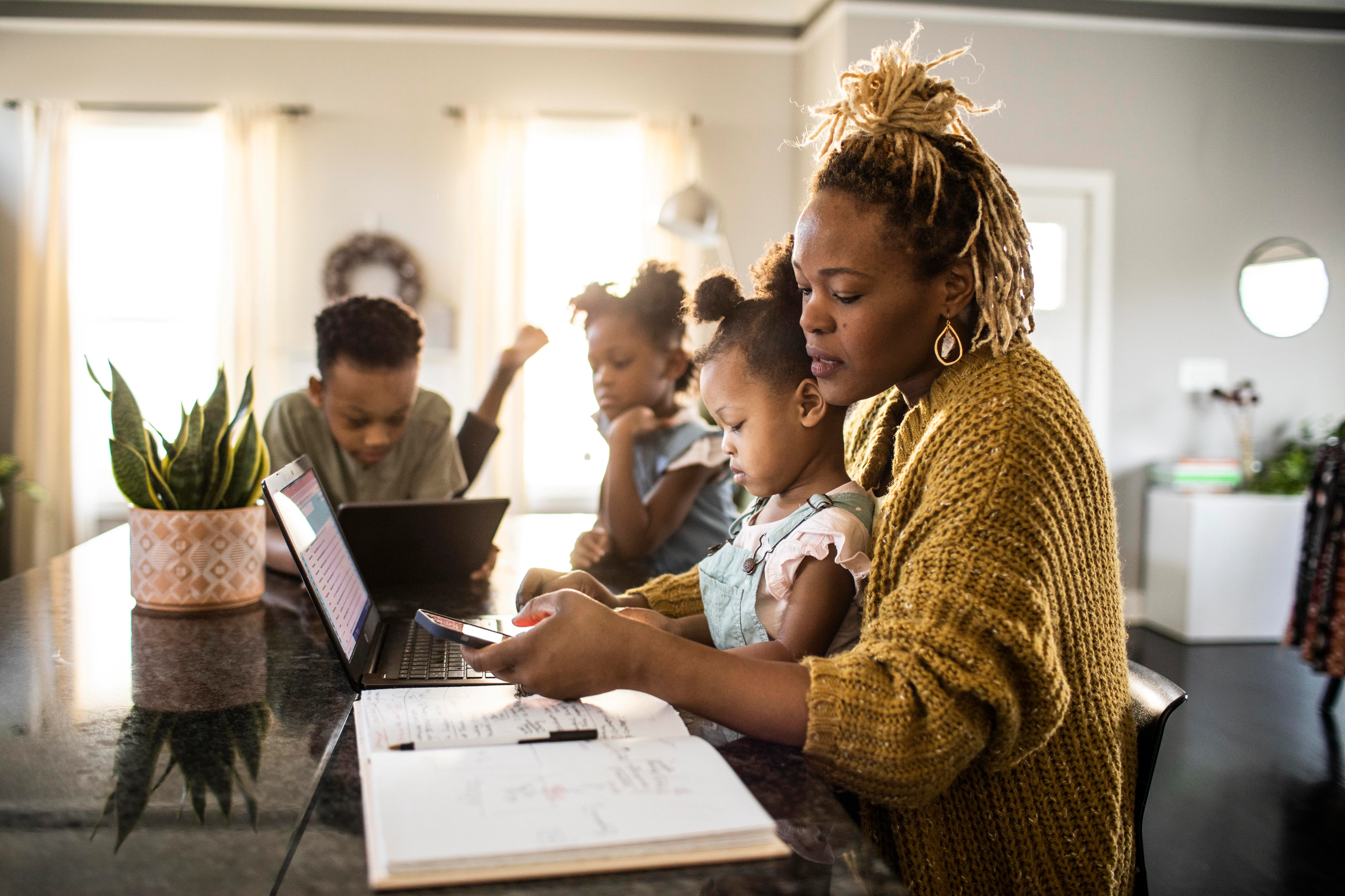 A mother helps her young daughter with homework at a kitchen island while two other children use lap...